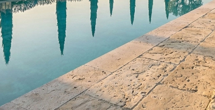 Vue rase sur plage de piscine en travertin vieilli — empreinte de pied mouillé sur la pierre, reflet figuier dans l'eau